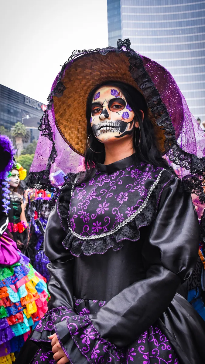 A woman in a wide-brimmed hat and black-and-purple dress with a painted skull face at a catrinas parade