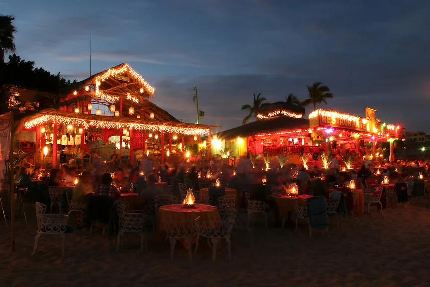 Mesas de restaurantes iluminadas de noche en la arena de Playa El Médano, Cabo San Lucas