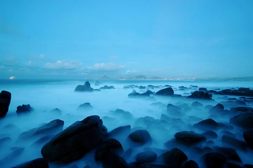 Olas sobre las rocas oscuras de Playa Monumentos al anochecer, con las luces de Cabo San Lucas a lo lejos
